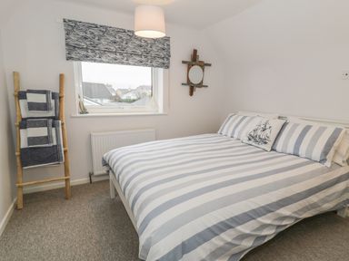 A bedroom with a double bed with striped bedding a window with patterned blind and a wooden towel ladder at Ty Taid in Rhosneigr