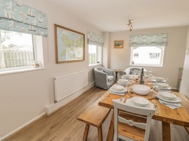 A dining room with a wooden table set for eight and two armchairs by windows with nautical curtains at Ty Taid in Rhosneigr