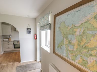 A hallway with a framed map on the wall a window with patterned blinds and a view into a kitchen at Ty Taid in Rhosneigr