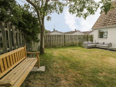 A backyard with a wooden bench under a tree and outdoor sofa seating near a white house at Ty Taid in Rhosneigr