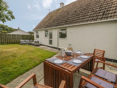A backyard with a wooden dining table set with plates and glasses and a patio sofa near a house at Ty Taid in Rhosneigr