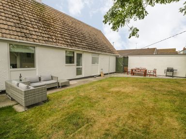 An outdoor patio area with wicker seating and a wooden dining set on grass at Ty Taid in Rhosneigr
