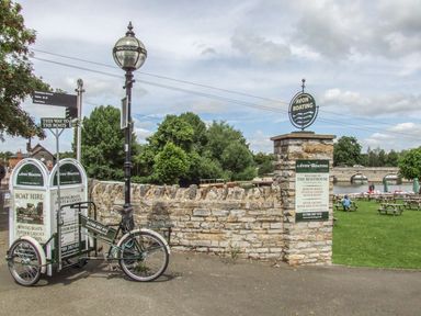 A riverside area with signs for Avon Boating a bike with boat hire signs and tables on grass by the river at Megs Cottage in Tiddington