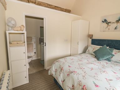 A bedroom with a bed with floral bedding a white wardrobe a shelving unit and an open door to an en suite bathroom at Megs Cottage in Tiddington