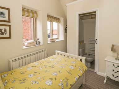 A bedroom with a bed with yellow floral bedding two windows with towels and a stuffed toy a white nightstand with a lamp and an open door to a bathroom at Megs Cottage in Tiddington