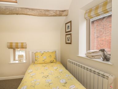A small bedroom with a single bed with yellow floral bedding two windows with yellow striped blinds and framed pictures on the wall at Megs Cottage in Tiddington