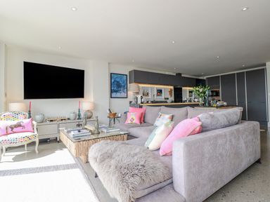 A living room with a large gray sofa with colorful pillows a patterned armchair a wooden coffee table and a kitchen area in the background at Stargazers in Sennen