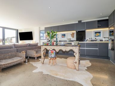 An open plan kitchen and living area with a gray sofa two wicker baskets an island table and a television at Stargazers in Sennen