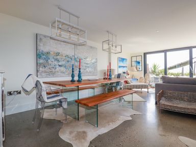 A dining room with a wooden table bench and chairs next to a living area with sofa and large windows at Stargazers in Sennen