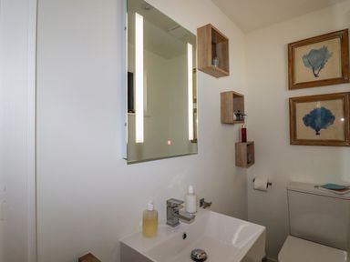 A bathroom with a sink and mirror above it and three wooden shelves on the wall with framed pictures above the toilet at Stargazers in Sennen