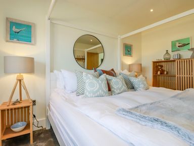 A bedroom with a white canopy bed with multiple pillows a wooden side table with a lamp a wooden cabinet with decorations and paintings on the walls at Stargazers in Sennen