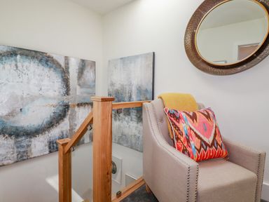 A corner with a beige armchair with a colorful pillow and yellow throw a round mirror on the wall wooden stair railing and abstract paintings at Stargazers in Sennen