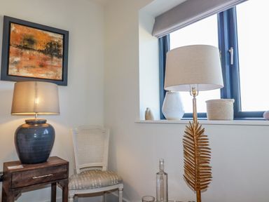 A corner of a room with a wooden side table and lamp a white chair a floor lamp and a window with decorations on the sill at Stargazers in Sennen