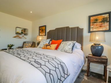 A bedroom with a bed with pillows and a blanket a bedside table with a lamp an armchair and wall art at Stargazers in Sennen