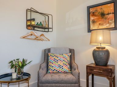 A corner with a gray armchair with a colorful pillow beside a small wooden table with a lamp and a round side table with a vase of flowers and a candle at Stargazers in Sennen