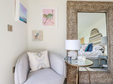A corner with a cushioned chair and a pillow next to a round side table with a lamp and decorative items in front of a large ornate mirror in a bedroom at Stargazers in Sennen