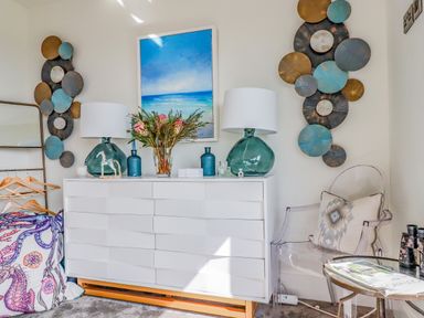 A white dresser with lamps vases flowers and a horse figurine a transparent chair with a pillow a small table with magazines and binoculars and wall art at Stargazers in Sennen