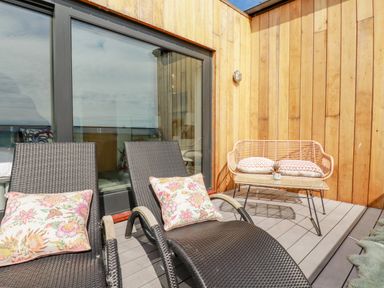A wooden deck with two brown lounge chairs with floral cushions and a small wicker bench with two pillows and a table at Stargazers in Sennen