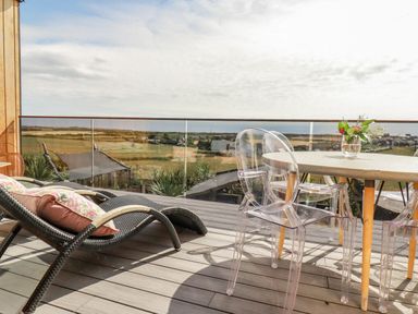 A balcony with wicker lounge chairs with cushions a round table with clear chairs and a vase with flowers overlooking fields and a village at Stargazers in Sennen