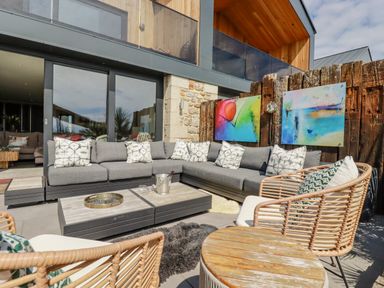 An outdoor seating area with a gray sectional sofa cushions two wicker chairs and two abstract paintings on a wooden fence at Stargazers in Sennen