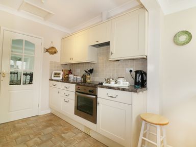 A kitchen with white cabinets oven kettle wall clock and a wooden stool at Sheilas Cottage in Penryn