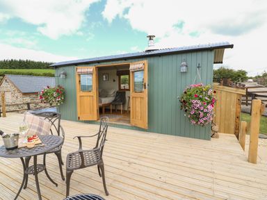 A wooden deck with metal table and chairs outside a green cabin with open wooden doors and hanging flower baskets at Ivys Way in Rhayader