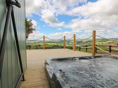 An outdoor deck with a hot tub and rope railing overlooking green hills and a cloudy sky at Ivys Way in Rhayader