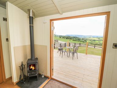 A room with a wood burning stove and a view of a wooden deck with a table and two chairs overlooking green fields at Ivys Way in Rhayader