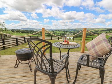 A wooden deck with metal chairs and a table with drinks and snacks overlooking green hills and fields at Ivys Way in Rhayader
