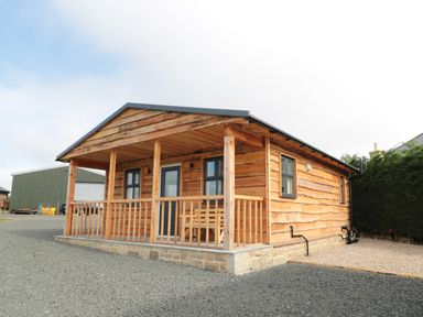 A wooden cabin with a porch and railing on a gravel lot at Aln in Longframlington