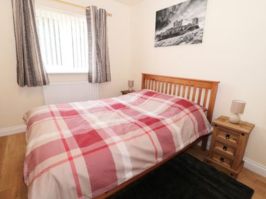A bedroom with a wooden double bed with red plaid bedding two wooden nightstands with lamps and a window with striped curtains at Breamish in Longframlington