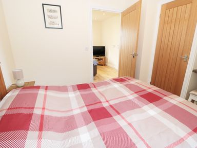 A bedroom with a bed with red and beige checkered bedding a bedside table with a lamp two wooden doors and a view to another room with a television at Breamish in Longframlington