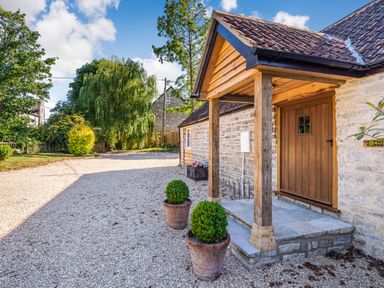 The entrance of a stone house with a wooden door covered by a porch and gravel driveway with potted plants at Old Farm Stables South Barrow near Sparkford