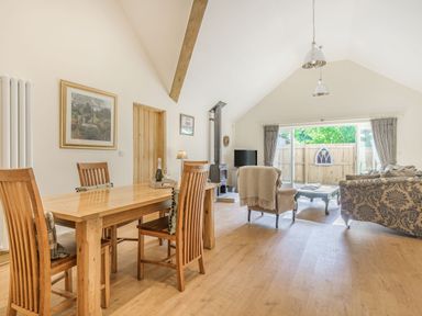 A dining area with wooden table and chairs next to a living space with armchairs, sofa, coffee table, stove, and large patio doors at Old Farm Stables South Barrow near Sparkford