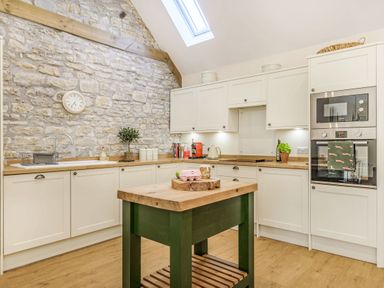 A kitchen with white cabinets a wooden countertop a stone wall a small island and built in oven at Old Farm Stables South Barrow near Sparkford