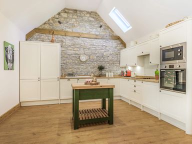 A kitchen with a stone wall and white cabinets with a small green island in the center at Old Farm Stables South Barrow near Sparkford