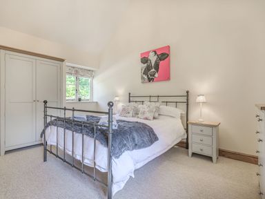 A bedroom with a metal bed frame white bedding a gray throw pillows a gray bedside table with a lamp a wardrobe and a window with a floral blind at Old Farm Stables South Barrow near Sparkford