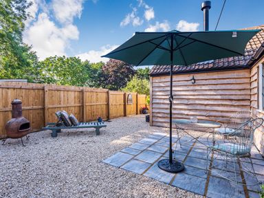 An outdoor patio area with a table and two chairs under an umbrella a wooden chaise lounge a fire pit and a wooden fence at Old Farm Stables South Barrow near Sparkford