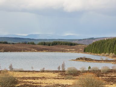A landscape with a lake surrounded by trees and hills under a cloudy sky at Burnside Lodge in Aberfeldy