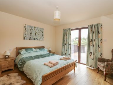 A bedroom with a wooden bed two bedside tables lamps a floral armchair and glass doors with curtains at Burnside Lodge in Aberfeldy