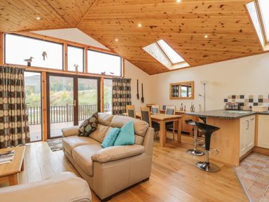 A living room with a beige leather sofa and blue cushions near dining table and kitchen bar at Burnside Lodge Aberfeldy