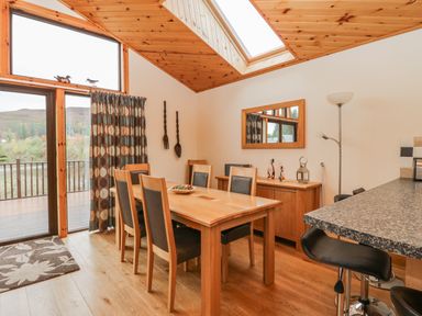 A dining area with a wooden table and six chairs near a sliding glass door with curtains and a skylight at Burnside Lodge in Aberfeldy
