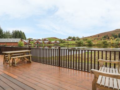 A wooden deck with a picnic table and bench overlooking a lake and wooden lodges in the countryside at Burnside Lodge in Aberfeldy