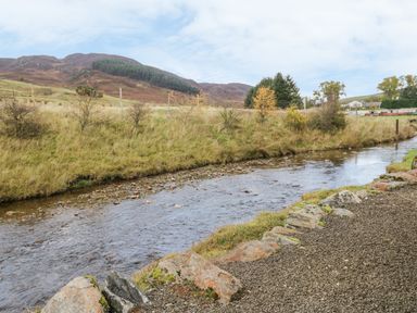 A river flowing beside a gravel path with hills and trees in the background at Burnside Lodge in Aberfeldy