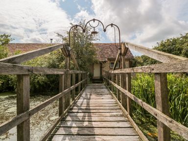 A wooden bridge over a pond leading to a stone building with a tiled roof at Jackdaw's Roost in Cheltenham
