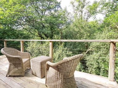 A wooden deck with two wicker chairs and a wicker table overlooking green trees at 4 Stonerush Valley in Lanreath
