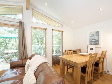 A dining area with a wooden table and chairs next to a brown leather sofa and large windows at 7 Streamside Lanreath