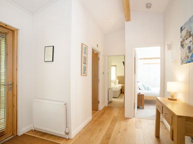 A hallway with wooden floors a wooden door a radiator two open doors leading to bedrooms and a wooden table with a lamp at 7 Streamside in Lanreath