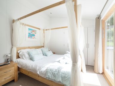 A bedroom with a wooden four-poster bed white bedding light blue pillows a wooden nightstand with a lamp a painting on the wall and a glass door at 7 Streamside in Lanreath