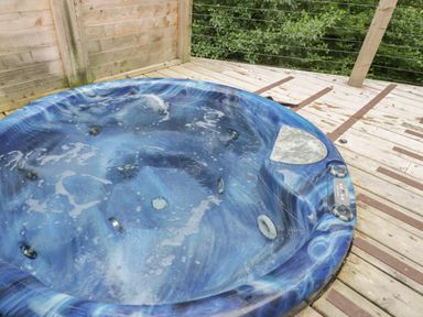 A blue outdoor hot tub on a wooden deck with railing and green trees in the background at 7 Streamside Lanreath